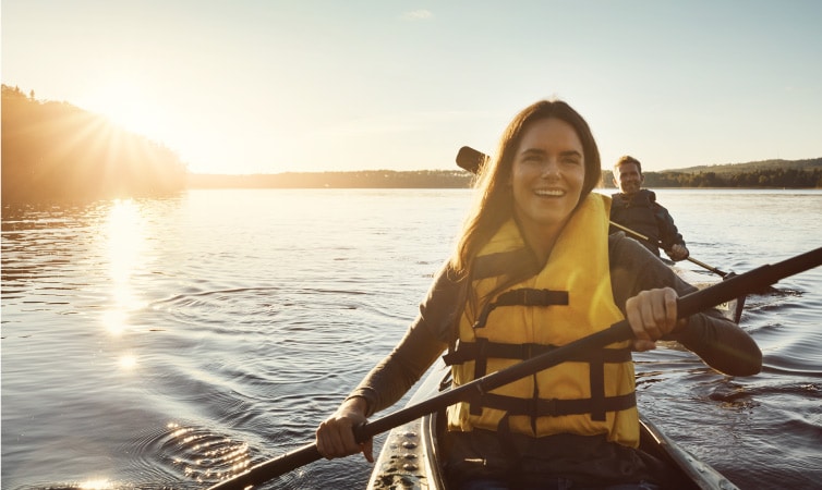 A happy couple kayaks on a peaceful lake as the sun rises over nearby trees.