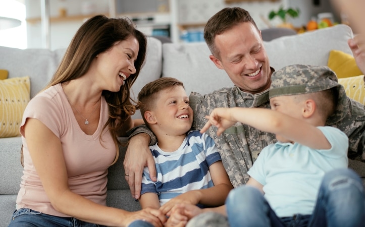 An American serviceman and his family are sitting on the floor in their living room, laughing together as one of his kids tries on his camouflage hat. 