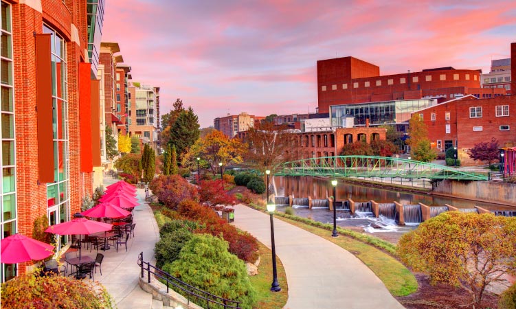 Red brick buildings stand along a Greenville, SC, waterway as the sun sets.
