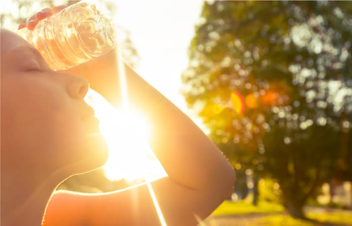 A young woman holds a water bottle against her forehead to cool herself down while the sun blazes in the background.