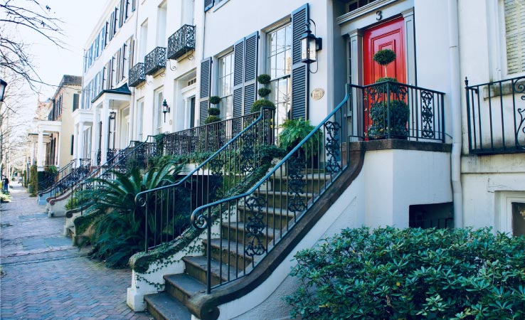 Historic townhome in Savannah, GA, with a red door, dark shutters, and wrought iron railing along the entryway stairs. 
