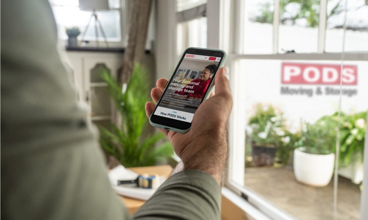 An arm is outstretched, and the hand is holding a smartphone with the PODS Boxes website pulled up. There is a PODS portable moving and storage container visible in the driveway through the window. Several potted plants are located inside and outside the home.