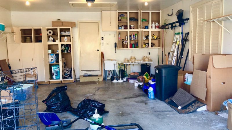 A "before" picture of a messy garage that's about to be decluttered. There are garbage bags and cardboard boxes on the floor, miscellaneous things are scattered about, and storage cabinet doors are left open.