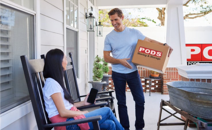 A man smiles at his wife while she sits on the porch of their new home in Savannah, GA. The man is holding a PODS moving box and there is a PODS moving container in their driveway. 