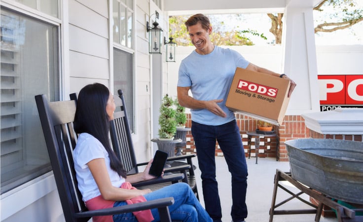 A man smiles at his wife while she sits on the porch of their new home in Savannah, GA. The man is holding a PODS moving box and there is a PODS moving container in their driveway.