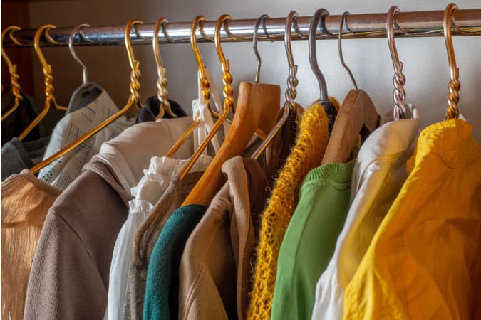 A dozen pieces of colorful clothing are hanging on a wardrobe bar. There are blouses and sweaters, and the hangers are gold and silver colored. There are a couple of wooden hangers, as well.