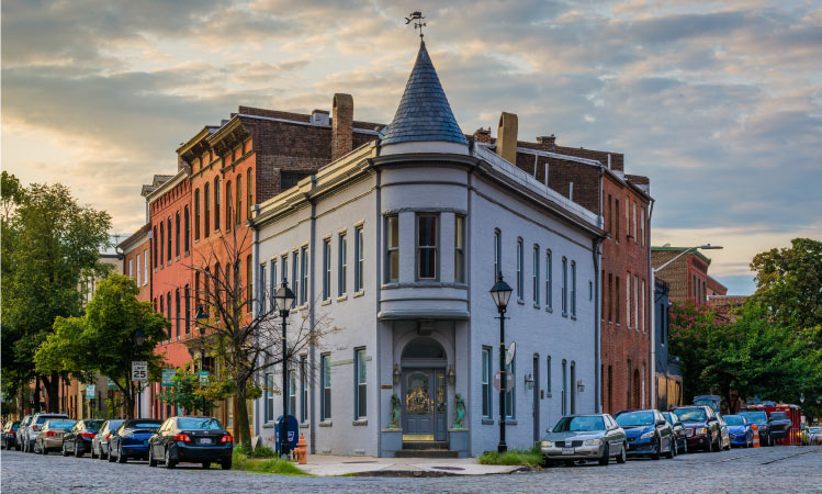 A perfect example of the historical architecture found at Fells Point is seen in a gray corner building with a spire on top. Both sides of the building are lined with cars, and other, more typical residential buildings are located further down the street.
