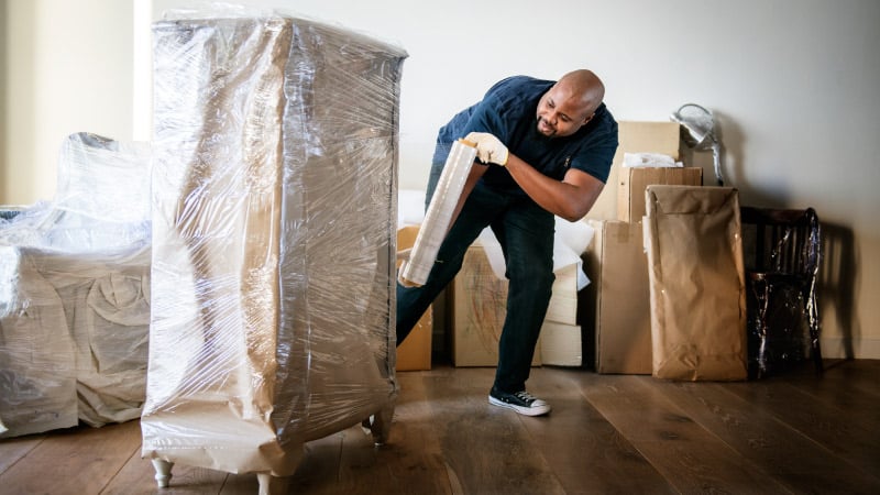 A man is wrapping a piece of furniture in plastic wrap before it is taken to long-term storage. Before wrapping it in plastic, the furniture piece was wrapped in packing paper. There are other pieces of wrapped furniture behind him.
