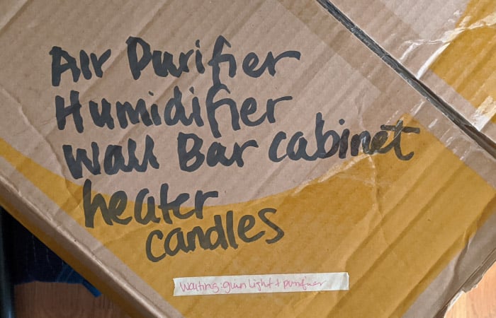 A moving box is labeled with “Air Purifier,” “Humidifier,” “Wall Bar Cabinet,” “Heater,” “Candles,” and a sticker that says it’s waiting on a light and purifier before being taped shut.