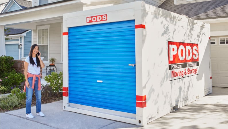 A woman talking on the phone in her driveway next to a PODS moving and storage container