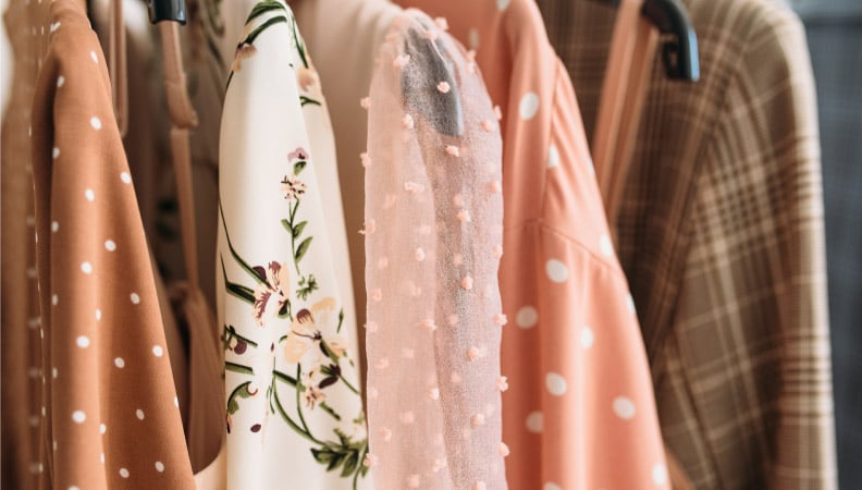 Several pretty blouses and sweaters hanging on a rack. They are light pink, white, and tan in color, and have various patterns and textures including floral, polka dot, and plaid.