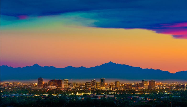 The Phoenix, Arizona, skyline at dusk. The distant mountains display a silhouette against the sky’s varying hues of yellow, orange, pink, and blue.