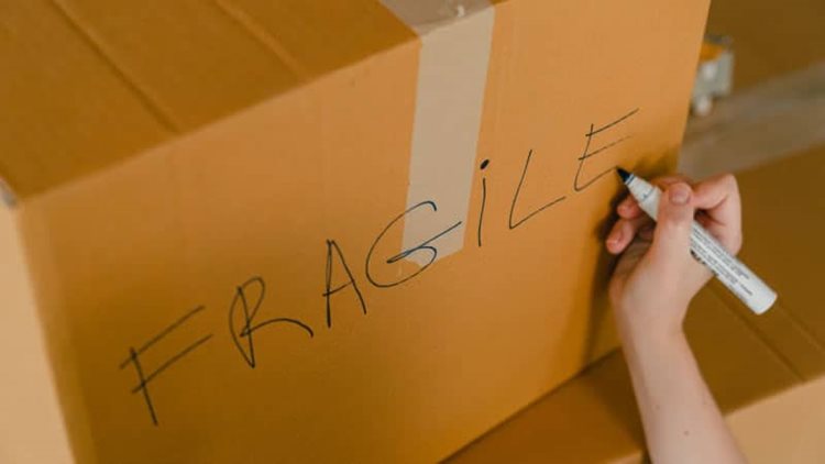 A woman's hand is writing the word "FRAGILE" on the side of a moving box that will be going into military storage.