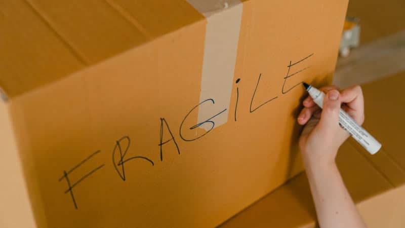 A woman's hand is writing the word "FRAGILE" on the side of a moving box that will be going into storage. 