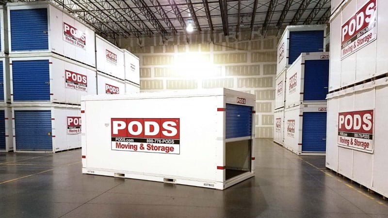 A PODS storage container is sitting on the floor of a secure PODS Storage Center with its door partially open. It is surrounded by several stacks of moving and storage containers.
