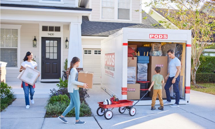 A family — consisting of a mother, daughter, son, and father — loading their PODS container in front of their old house. The mother is carrying a wrapped picture frame, the daughter is carrying a small box, and the son is pulling a red wagon to the father with items in the back of it. 