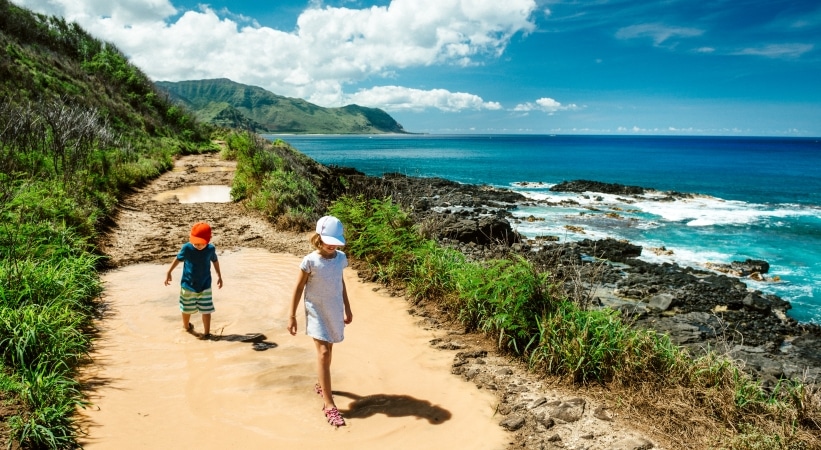 Two young children are walking along a coastal hiking path on Oahu. It's a sunny day and both the sky and sea are a beautiful blue color. 