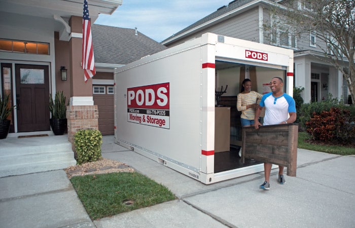 A man and woman are happily unloading their PODS portable moving and storage container in the driveway of their new house. The man is carrying a headboard, and the woman is carrying a box.