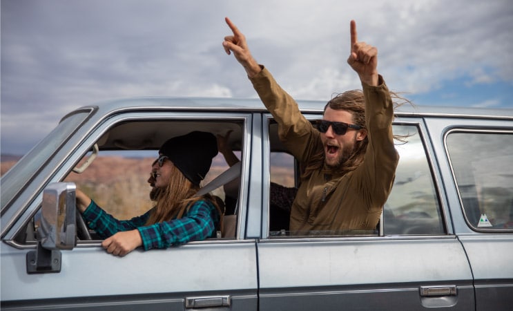 A group of friends is driving. One of the men is holding his arms out the window and shouting in excitement because he and his friends are moving to Texas.