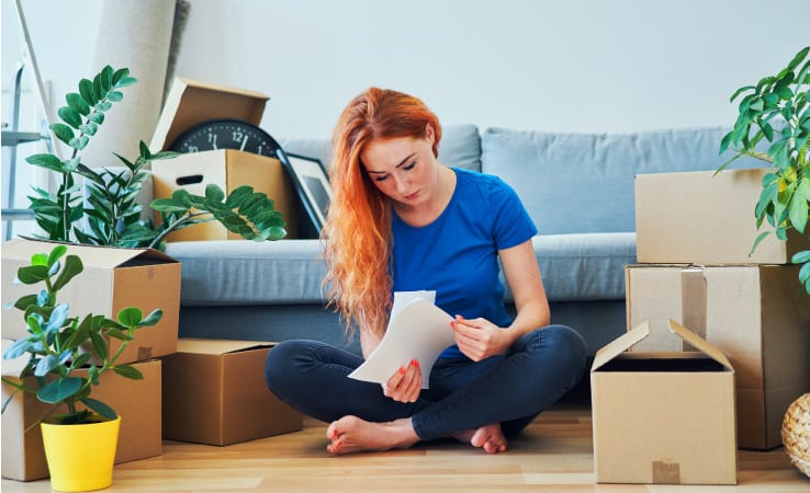 A young woman sits in her living room beside packed moving boxes and plants while she looks through moving quote.