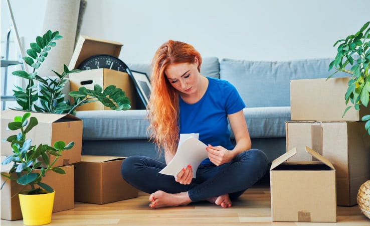 A young woman sits in her living room beside packed moving boxes and plants while she looks through moving quote. 