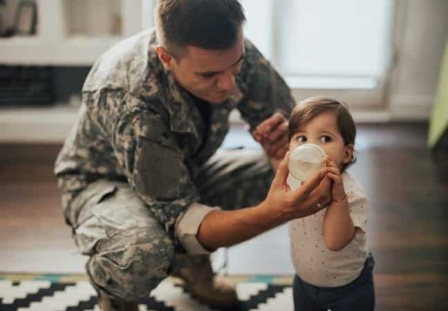 A U.S. serviceman in uniform is helping his baby drink from a bottle. 