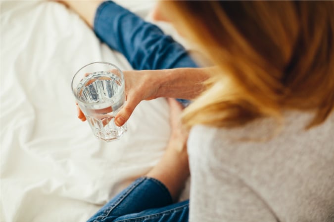 An over-the-shoulder view of a woman taking a break with a glass of water in hand. She has copper-colored hair and is wearing a gray knit top and blue jeans. She’s sitting on her bed with one leg bent and the other extended in front of her.