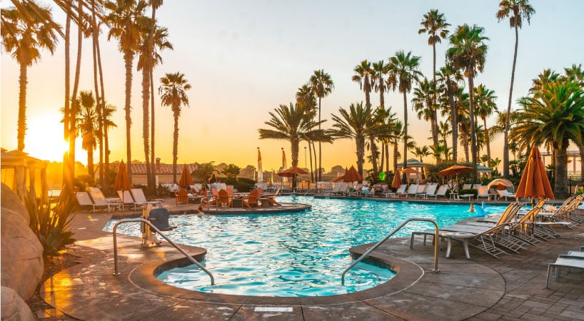 This resort-style pool is surrounded by chairs, palm trees, and a beautiful view of the sunset in the distance.