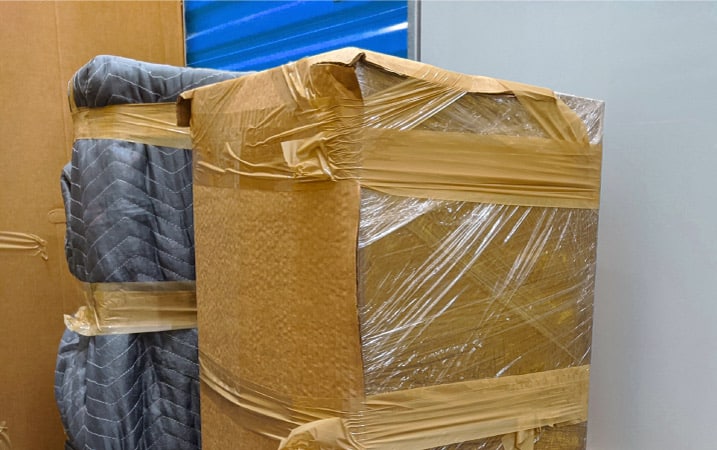 Two pieces of large furniture are wrapped in repurposed cardboard, moving blankets, and plastic wrap to protect them during a move.