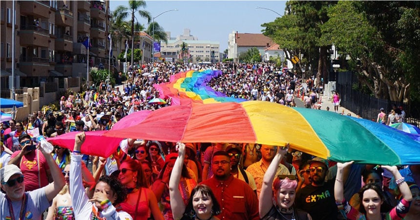 There is a very large parade going on, with people all along the way holding up a rainbow Pride flag  to celebrate LGBTQ+ awareness and support.