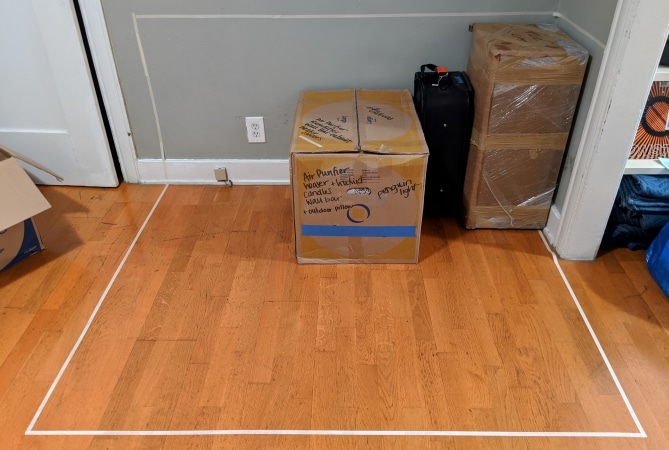 A corner of the room has been taped off to represent a moving container. Someone has stacked some boxes in this space to see how they will fit.