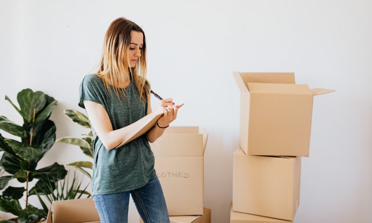 A young woman is standing beside stacks of assembled moving boxes and a few house plants. She’s holding a large notepad in her hand and checking things off a list.