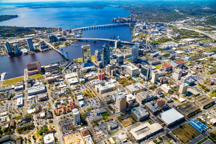 Aerial view of Jacksonville, Florida, where three of the city’s bridges cross the St. Johns River. The river waters are a dark blue and the city is a mix of greenspaces and city buildings of all sizes. 