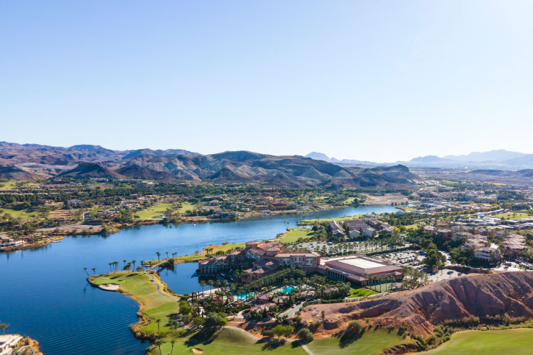 Aerial view of the Lake Las Vegas community in Henderson, Nevada, on a sunny day. The community features single-family homes and apartment buildings around a man-made lake, with golf courses and other amenities. 