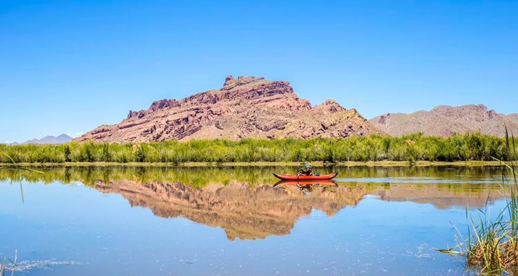 A lone kayaker glides down a glass-like river in Mesa, Arizona. The banks of the river are filled with tall grasses and, in the distance, large rock formations jut up into a clear blue sky. The water reflects the kayaker and the natural surroundings like a mirror. 