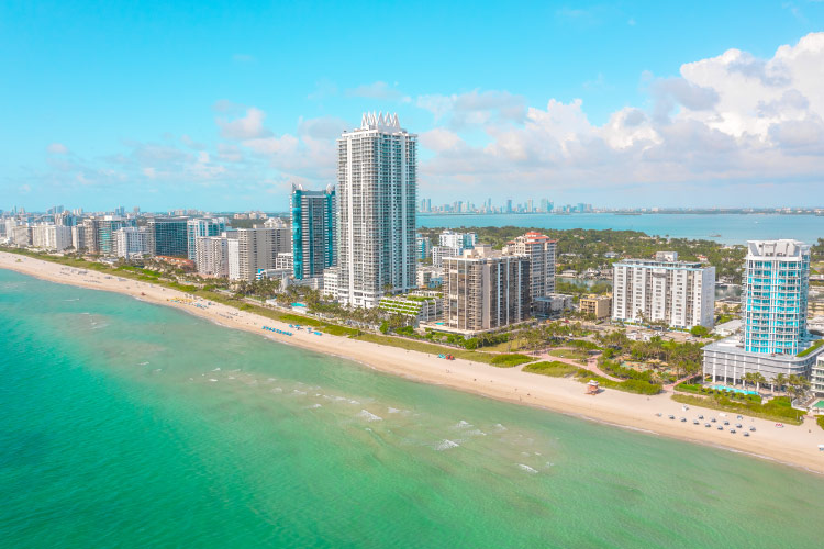 Aerial view of Miami Beach wih its turquoise waters lapping against sugar-sand beaches. Highrise condo buildings dot the shore and, in the distance, Downtown Miami can be seen. 