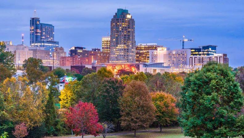 Dusk view of Downtown Raleigh buildings from Dorothea Dix Park in the fall. The sky is a dim blue and the city’s lights have begun to come on. Trees in the park are starting to change colors, fro green to red. 