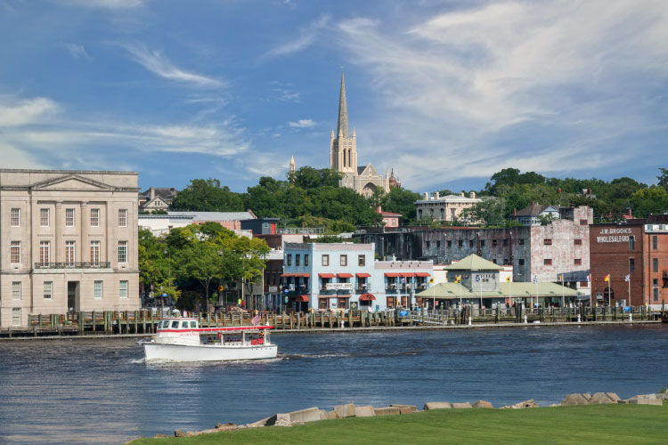 View of Wilmington, North Carolina, from across the Cape Fear River, on a summer day. A white boat motors past the riverfront buildings,and a tall steeple peeks out from above the mature trees in the city.