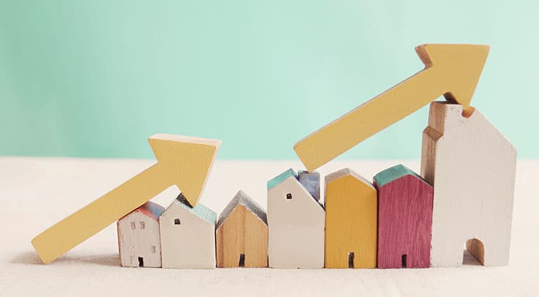 Small wooden carvings shaped like houses of various styles and colors are lined up from smallest to biggest. There are two wooden arrows leaning on the houses pointing upward to indicate an increase in housing costs.