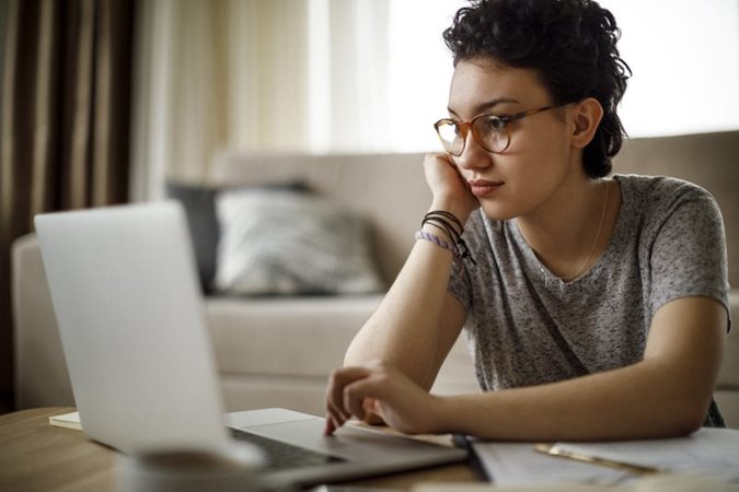 A woman is sitting in her living room at the coffee table, using her laptop to browse real estate and rental websites online before moving cross-country.