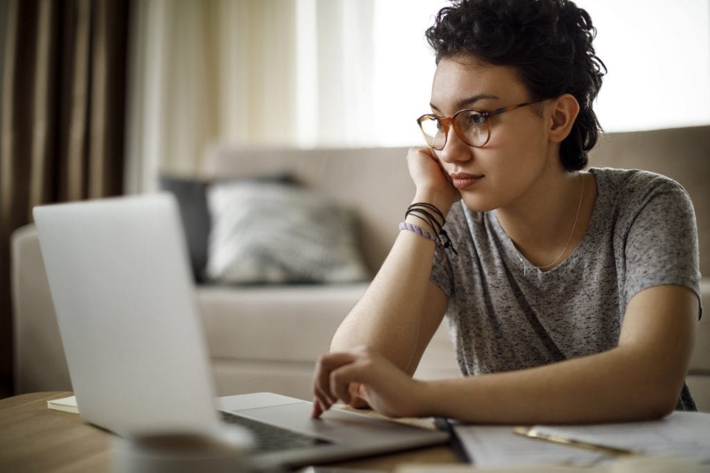 A woman is sitting in her living room at the coffee table, using her laptop to browse real estate and rental websites online. She is searching for housing before moving cross-country.