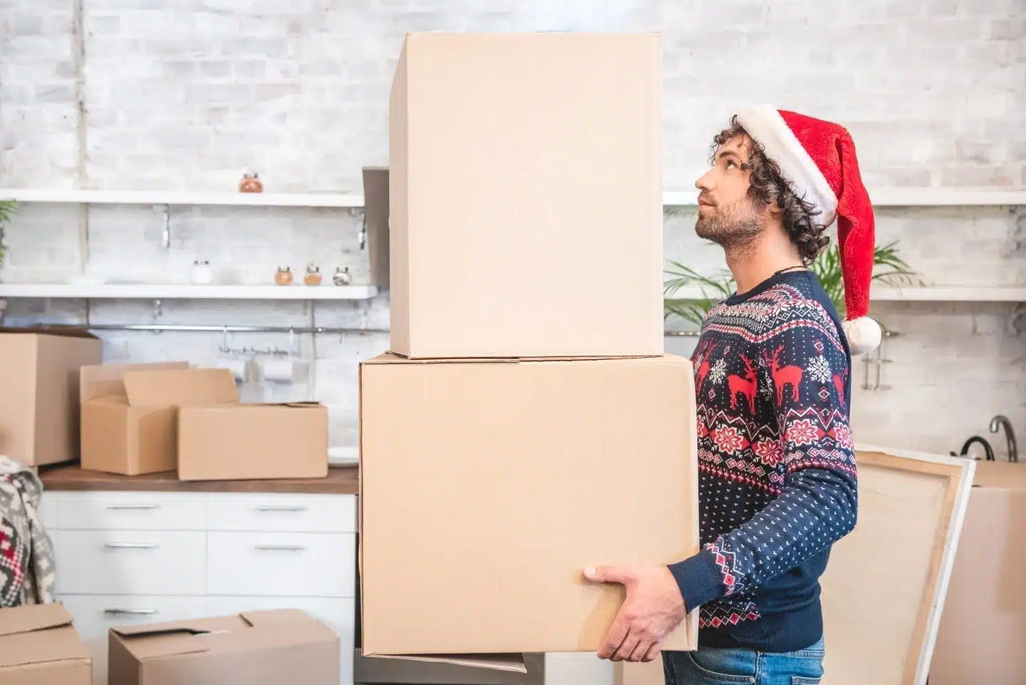A man wearing a holiday sweater and Santa hat stands in his kitchen, holding two large moving boxes. There are several more boxes of various sizes in the kitchen, making it clear that he’s moving during the holiday season.