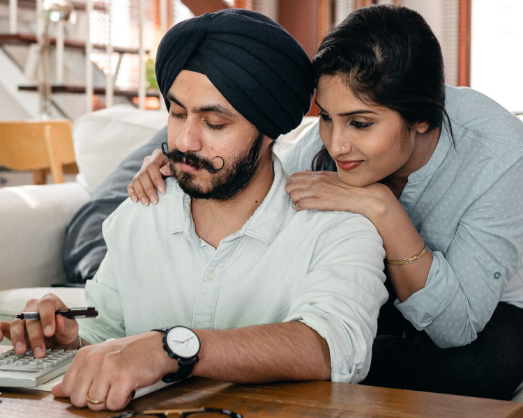 A woman affectionately leans against her husband’s back, watching as he uses a calculator to figure out their moving budget so they can decide when to move.