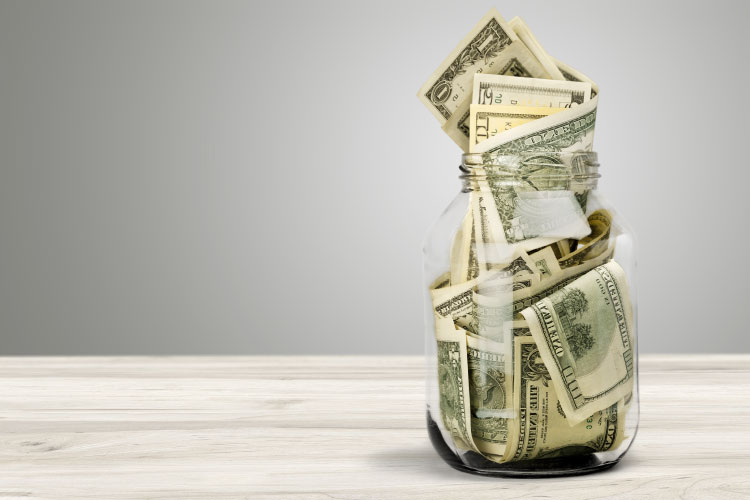 A glass jar stuffed full of cash savings sits on a light gray wooden table.