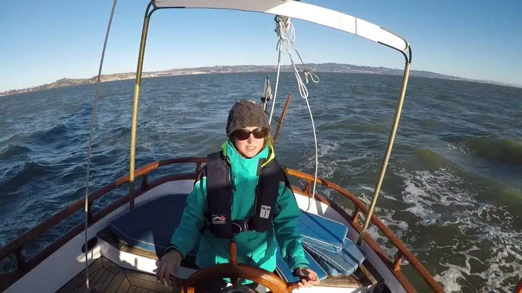 A woman dressed in cold-weather gear and a life jacket is steering her sailboat.