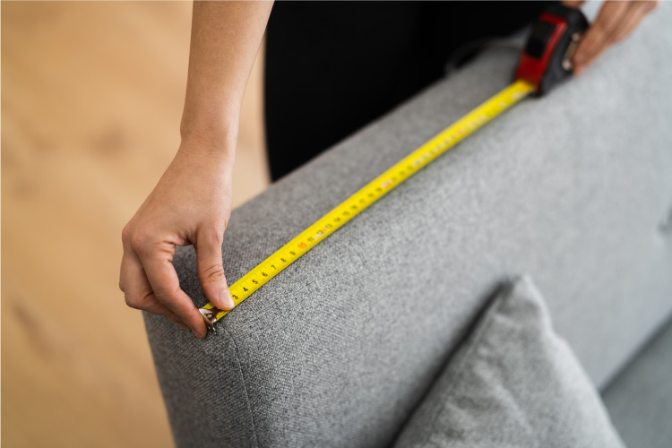 Close-up view of a pair of hands using a measuring tape to measure the armrest on a couch.