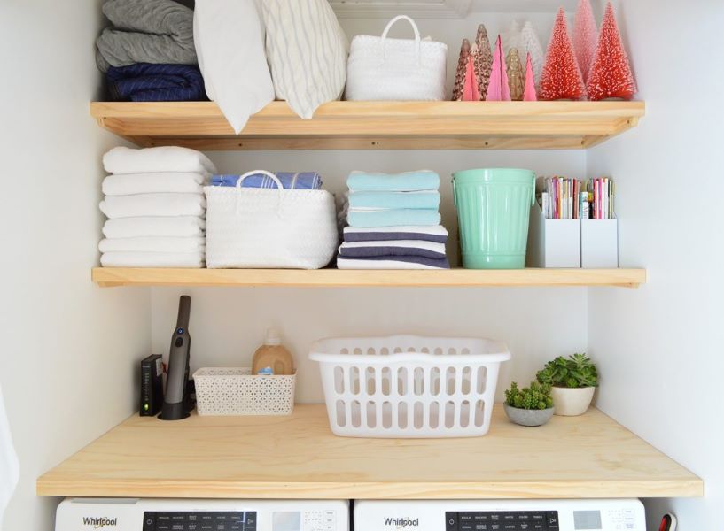 Neatly organized shelves above a residential washer and dryer are filled with towels, pillows, and other linens, as well as some cleaning supplies, art supplies, and seasonal decor.