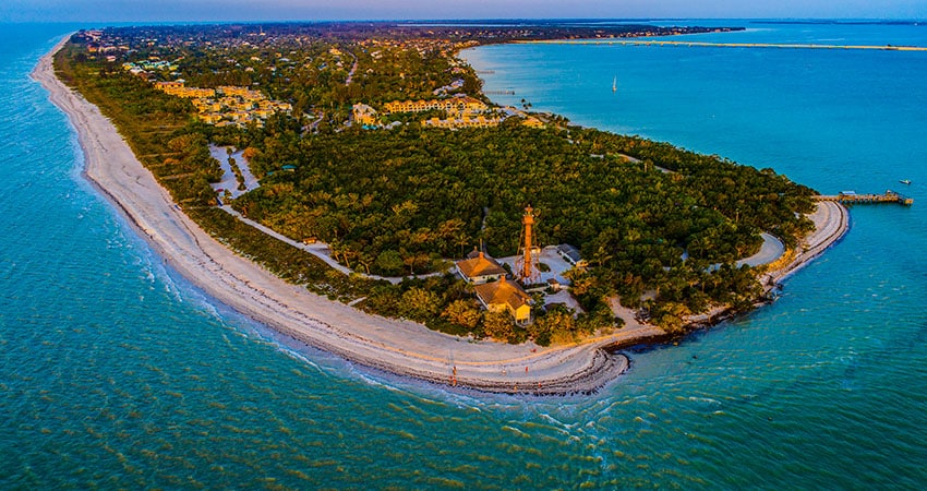 An aerial photo of Sanibel Island at sunset. The sun reflects off Sanibel's lighthouse and beach homes, bathing them in a golden glow. 