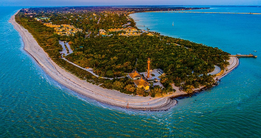 An aerial photo of Sanibel Island at sunset. The sun reflects off Sanibel's lighthouse and beach homes, bathing them in a golden glow.