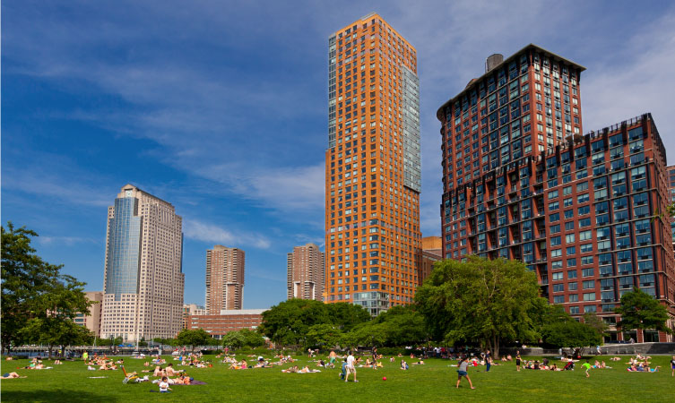 Dozens of locals enjoy a sunny day in a park in Battery Park City, New York, with residential high rises in the background. 